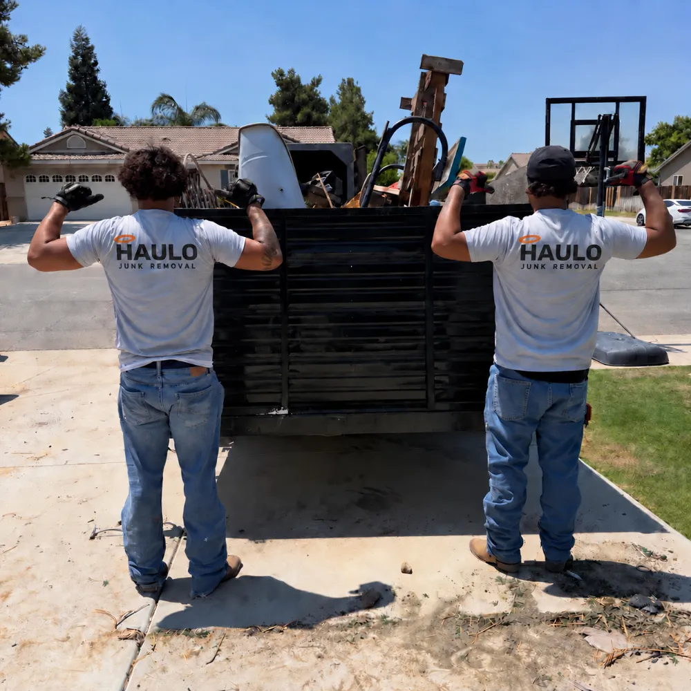 Two Haulo crew members in Haulo Junk Removal shirts at the back of the loaded dump trailer on a driveway