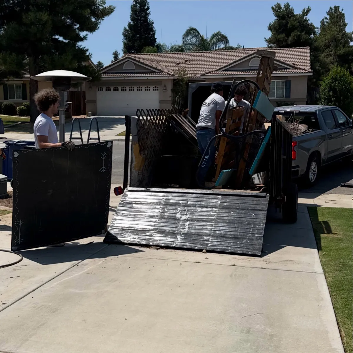 The Haulo crew loading lattice panels, boards and sheet metal into the dump trailer on a residential driveway, with the pickup truck hitched in front