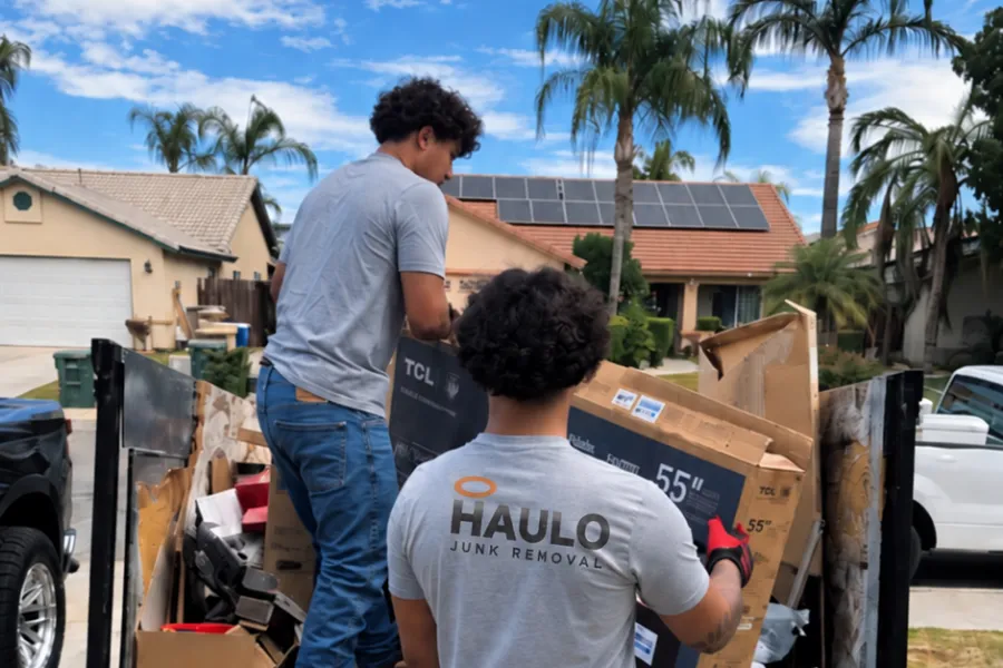 Two Haulo crew members loading flattened boxes and a TV box onto the dump trailer in a residential driveway