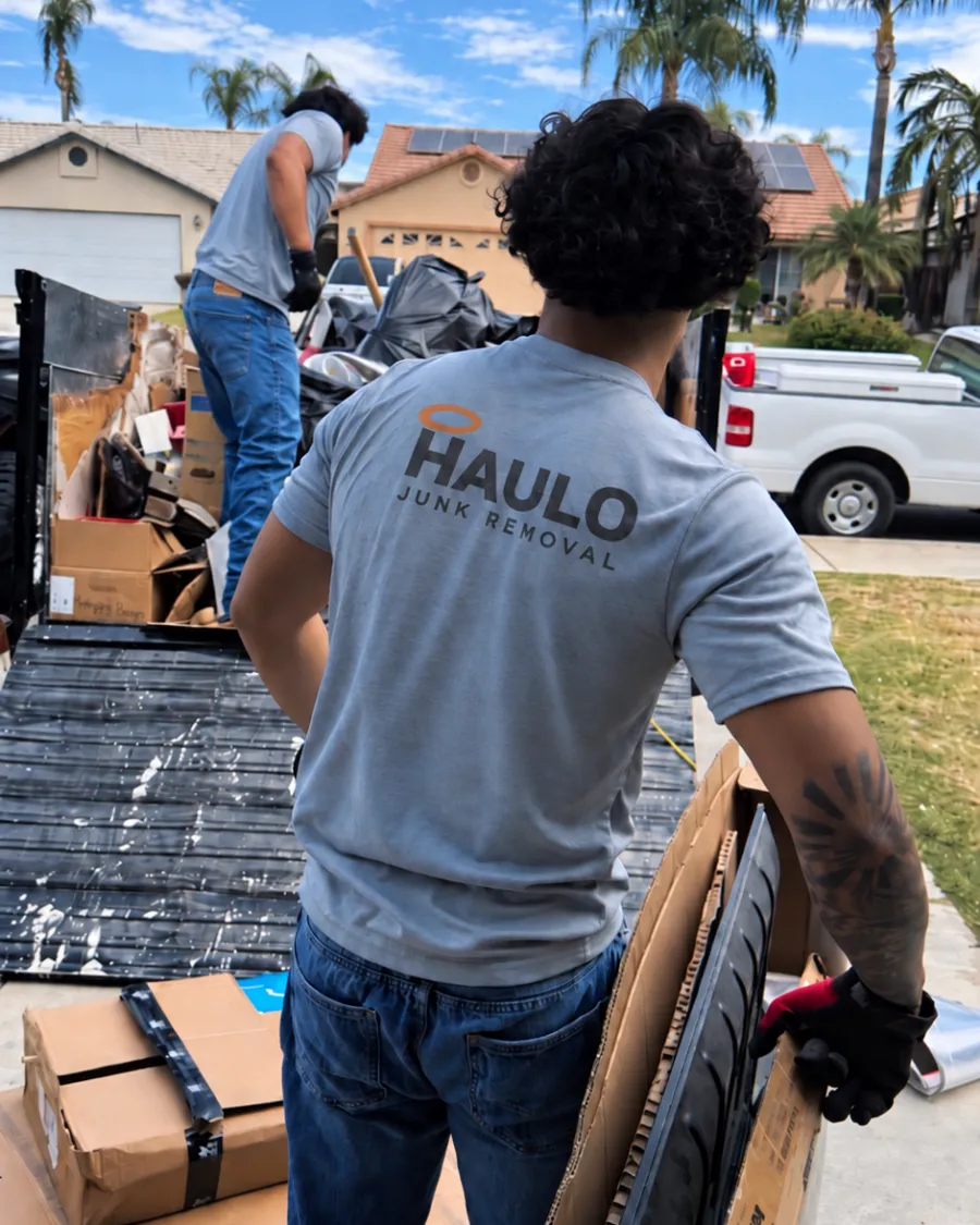 Haulo crew member in a Haulo Junk Removal shirt carrying a flattened TV box toward the loaded trailer while a second crew member stacks the load