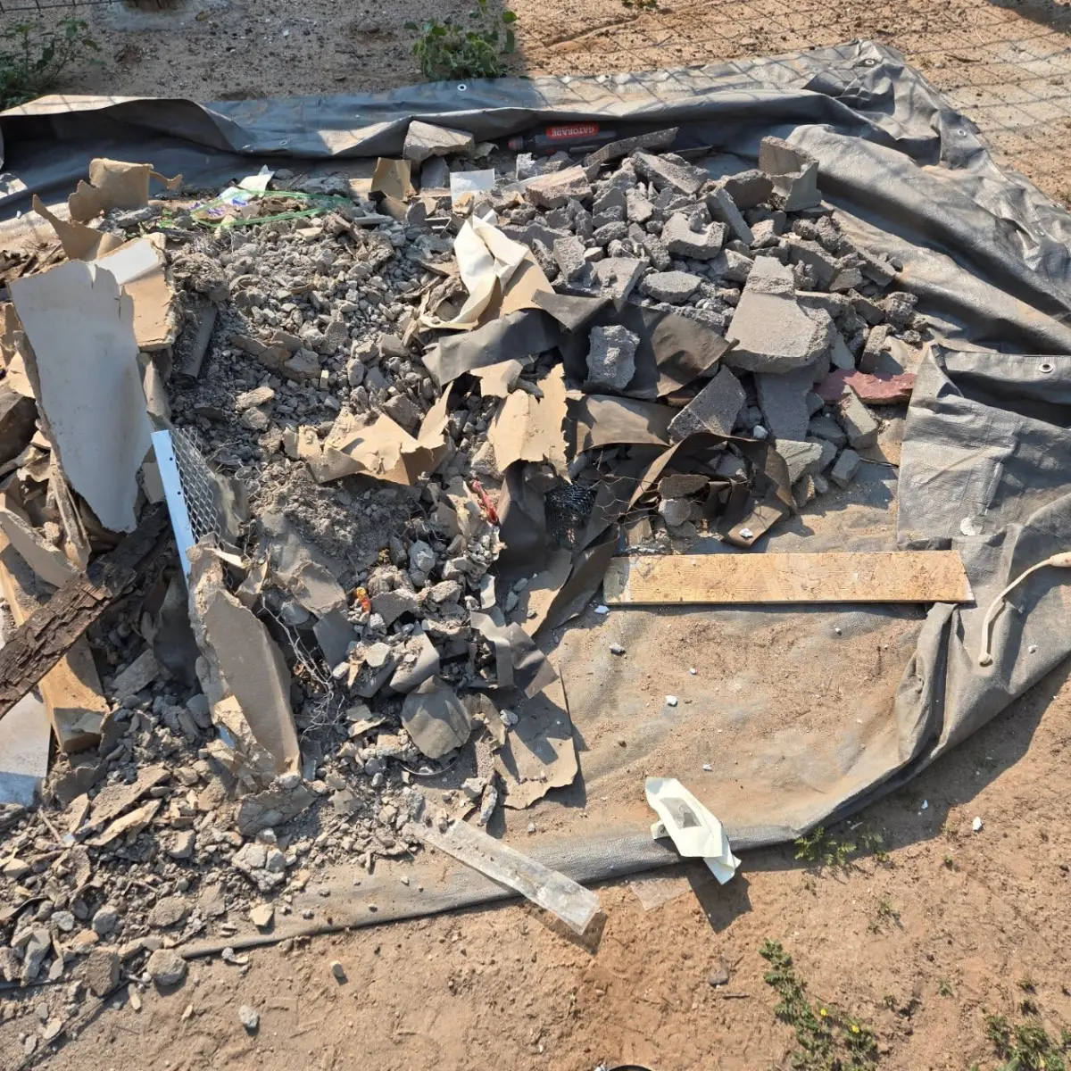 A pile of broken concrete and block rubble, drywall, stucco lath, wire mesh and wood offcuts on a grey tarp on bare dirt beside a wire fence in Apache Junction
