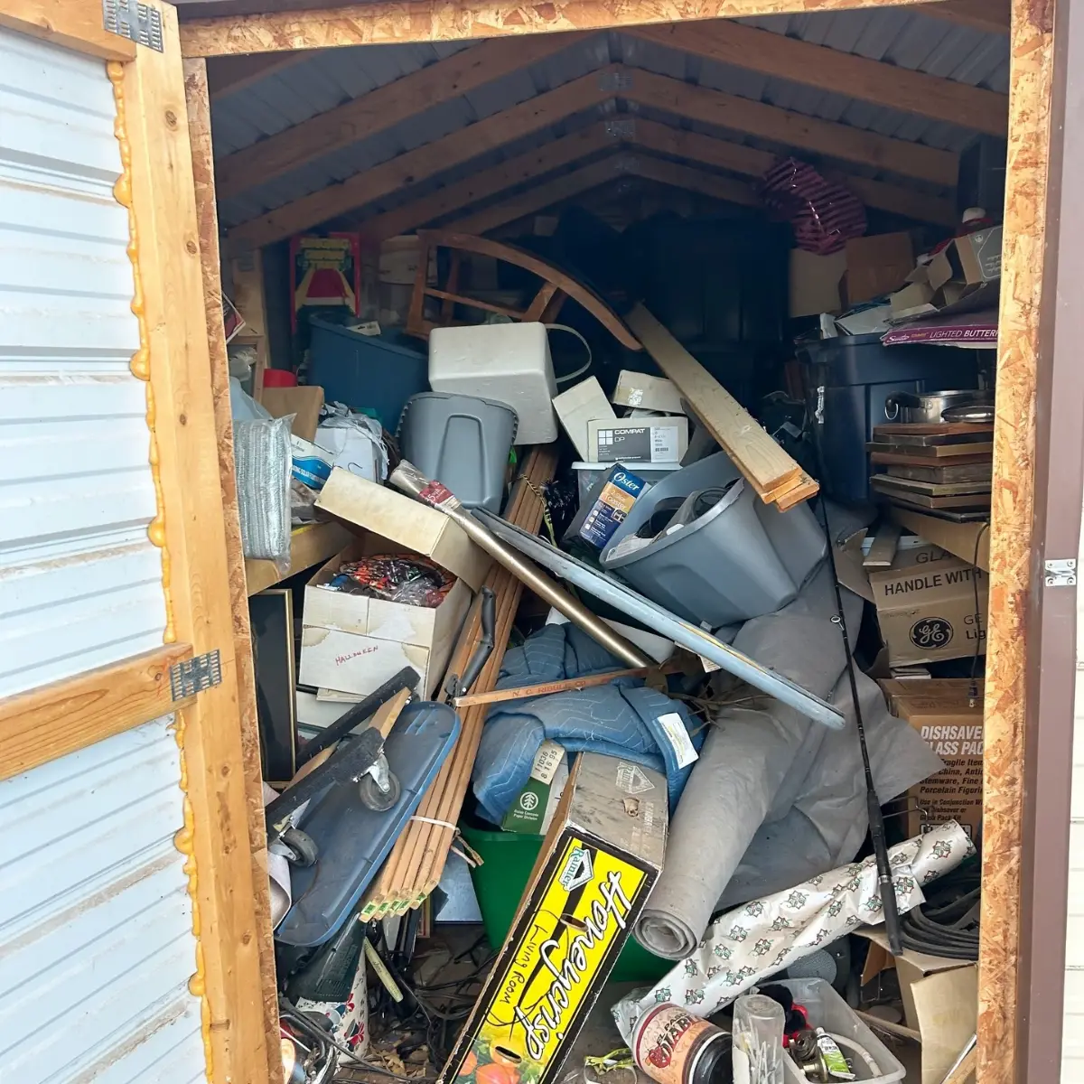 A metal-sided storage shed in Apache Junction packed floor to roof with plastic bins, boxes, moving blankets, a folding chair and loose contents