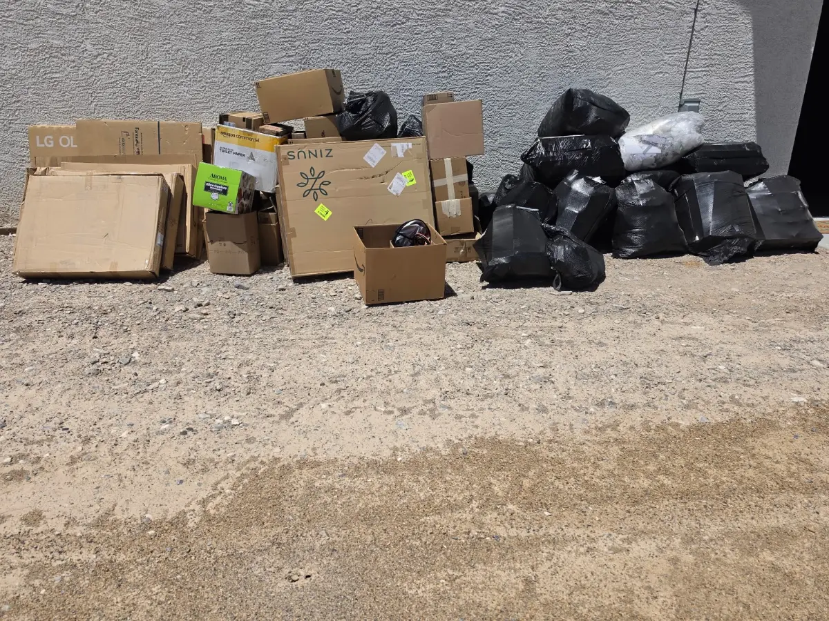 Cardboard boxes and bagged household trash piled against a stucco wall on a dirt side lot in Carefree