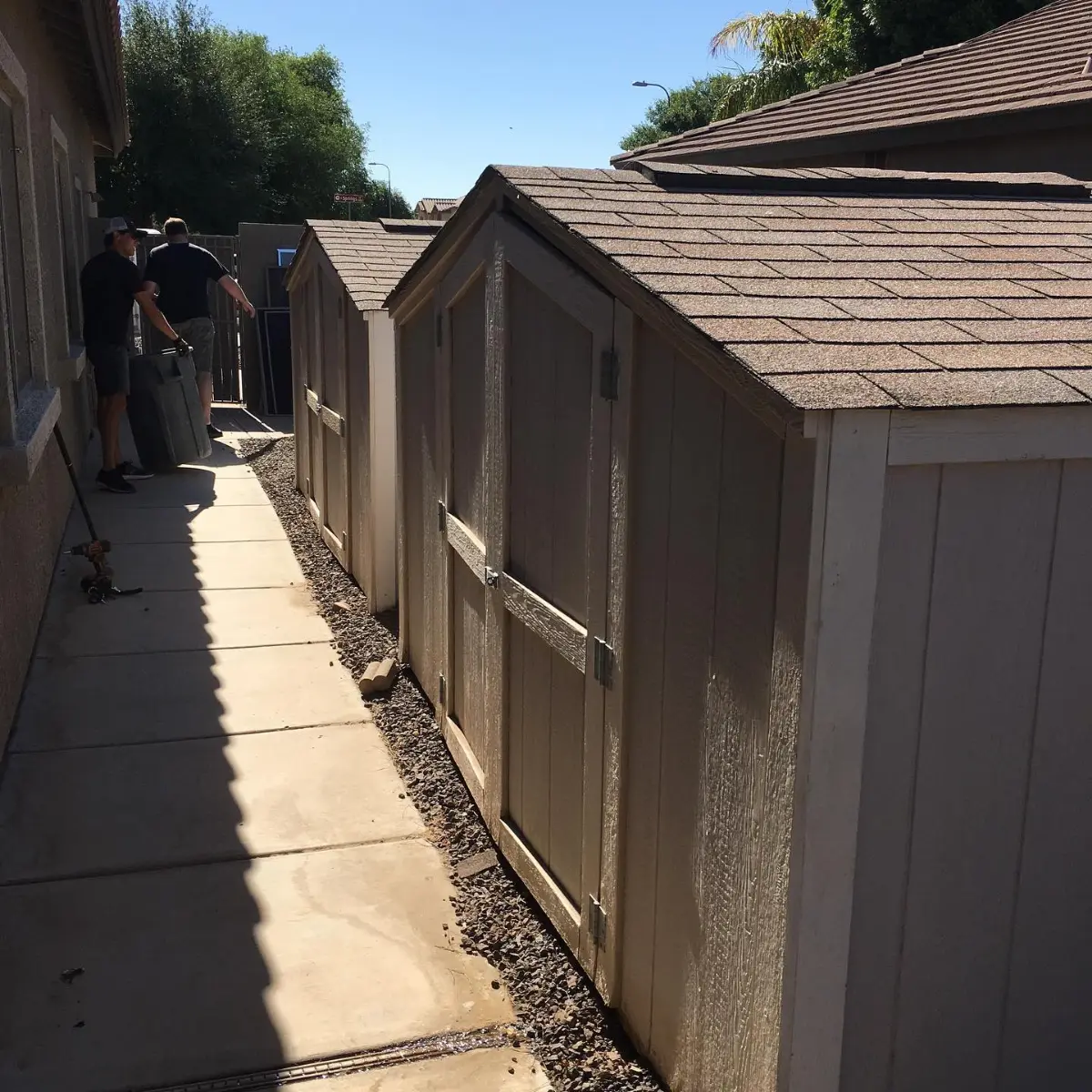 Three tan wood sheds with shingled roofs standing end to end along a narrow Chandler side yard, with two Haulo crew members at the side gate