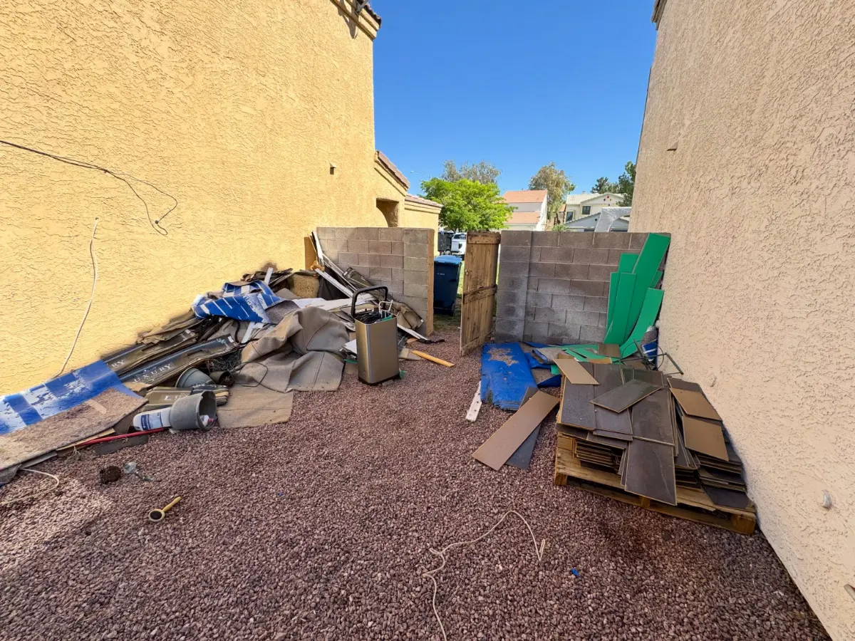 Carpet, padding, tile and flooring offcuts piled in a narrow gated side yard on red gravel in Gilbert