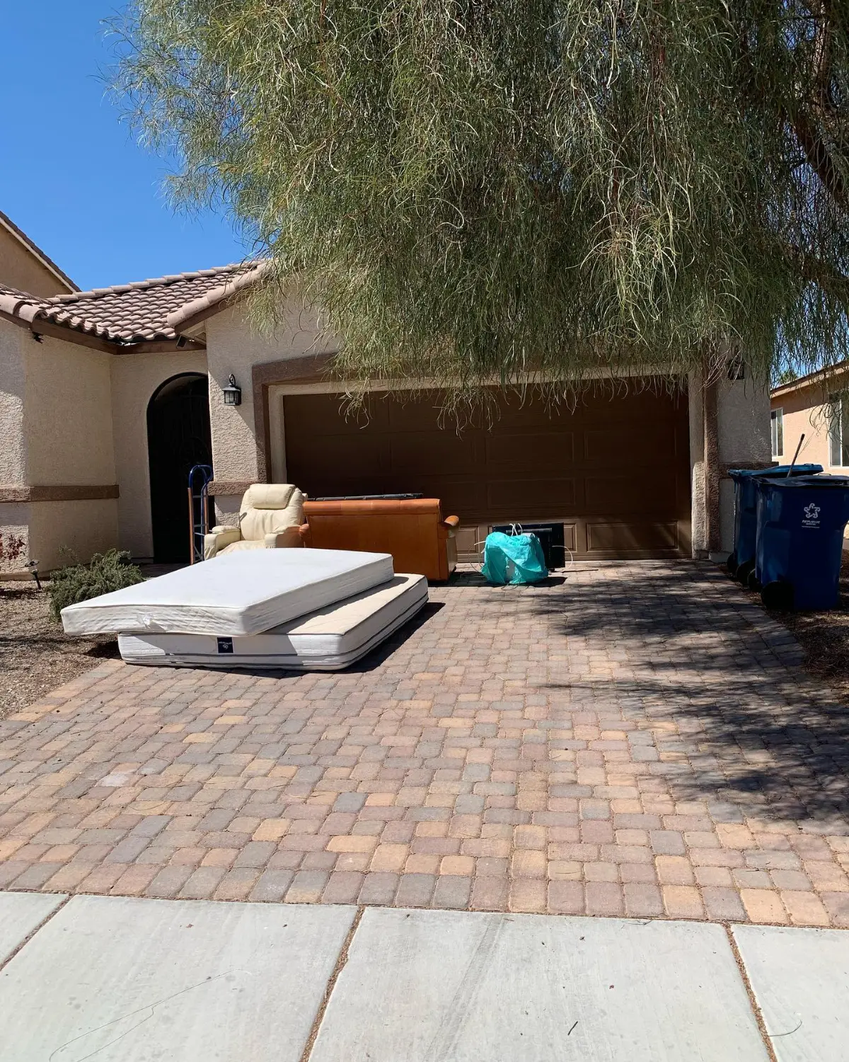 Two mattresses stacked on a Gilbert paver driveway beside a tan leather recliner and an orange leather sofa in front of a brown garage door under a large tree