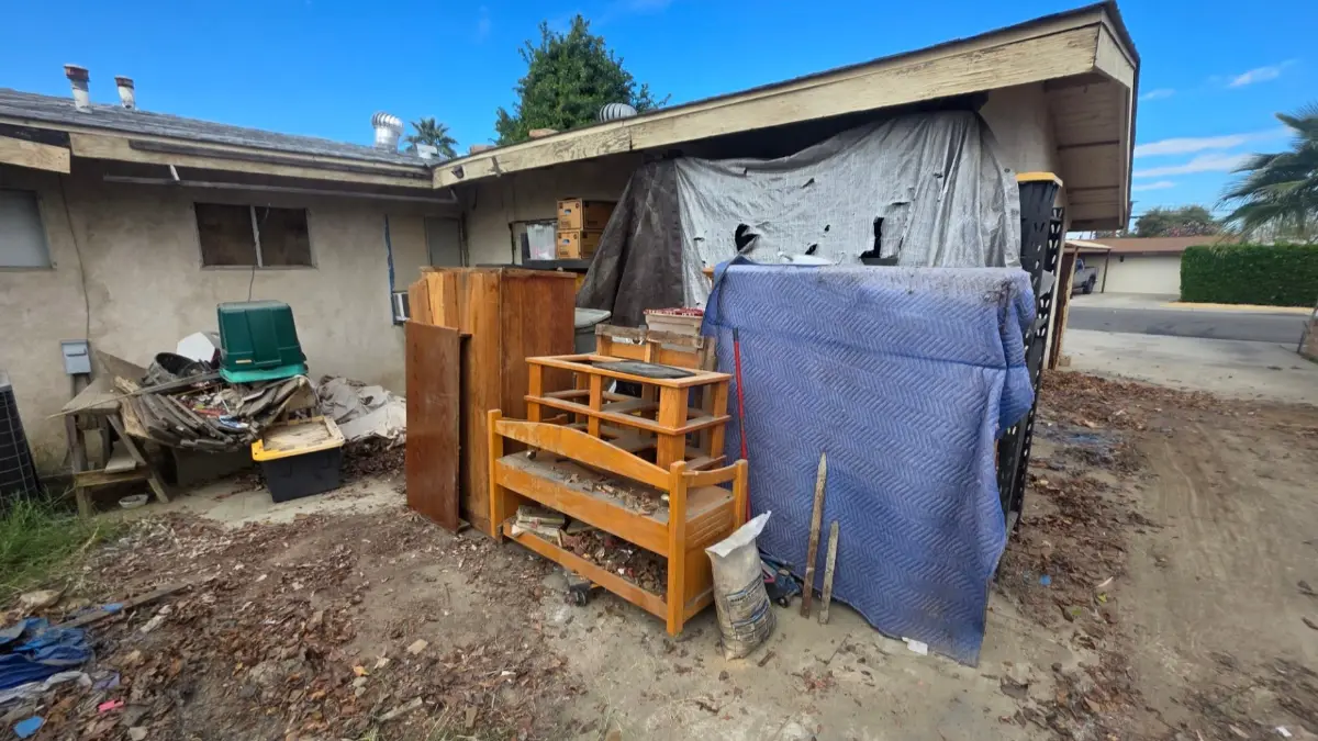 A cabinet, a wood bed frame, crates and a blue moving blanket under a side-yard carport at a Goodyear home