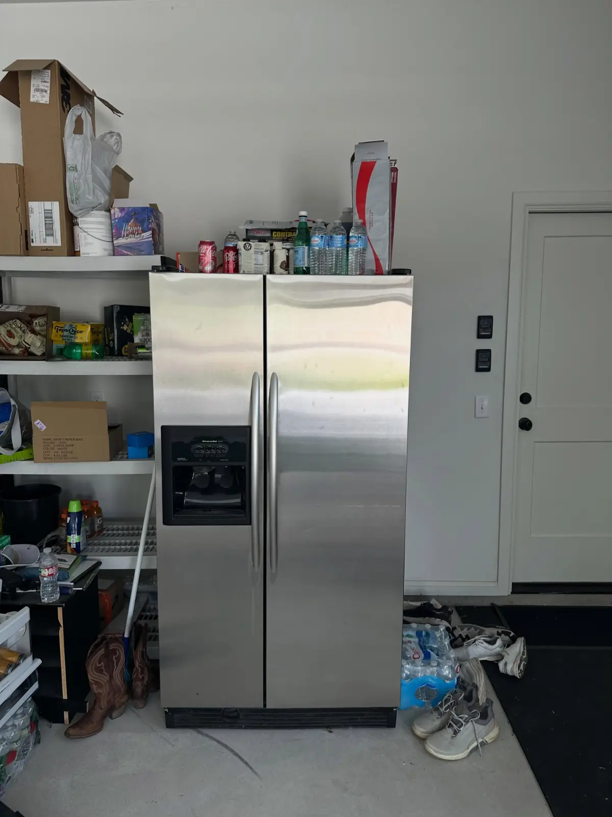 Stainless side-by-side refrigerator standing against a Mesa garage wall between shelving and a door