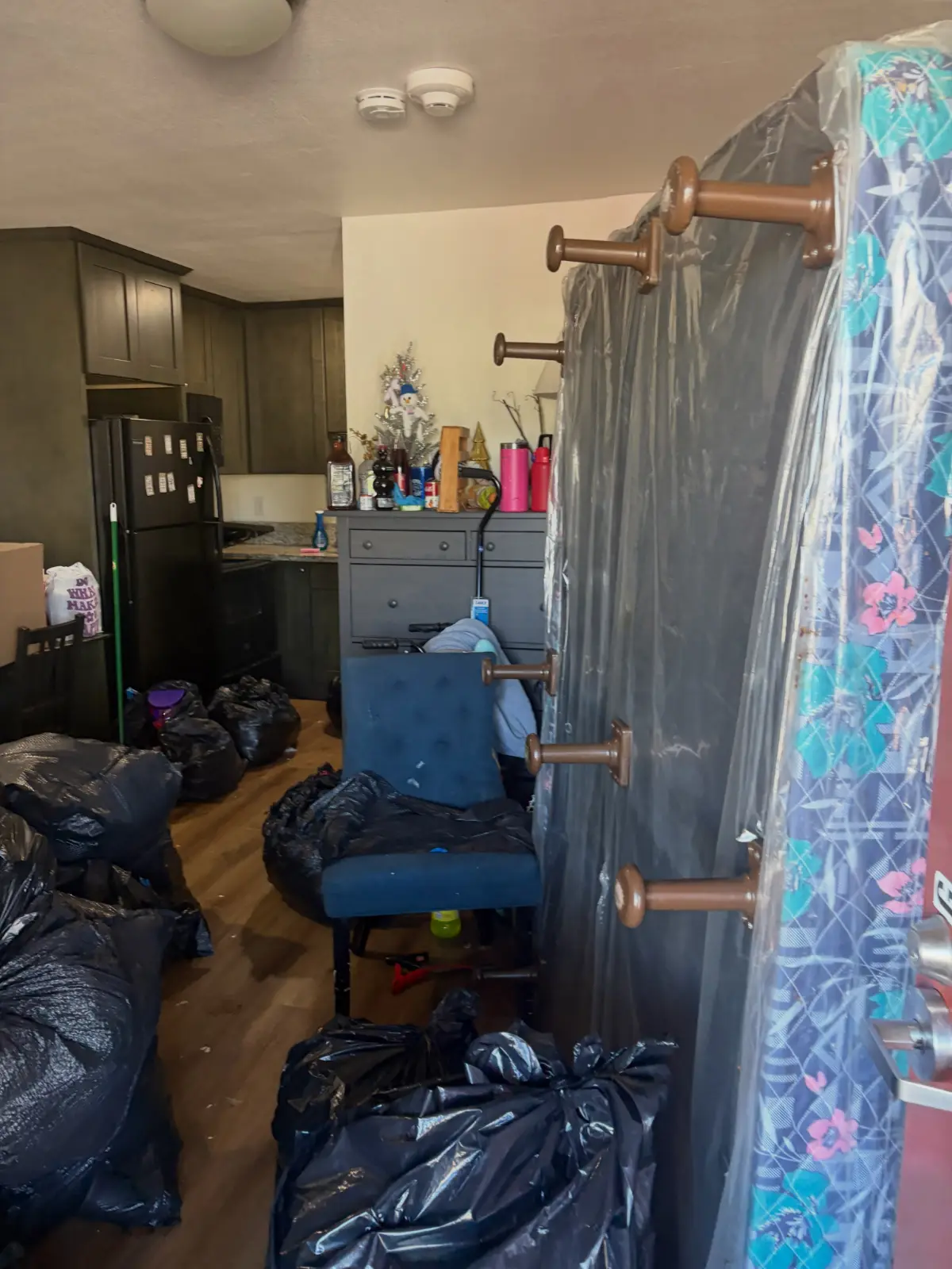 Kitchen and entry of a Mesa apartment unit filled with black trash bags, a plastic-wrapped mattress, a coat rack and an upholstered chair