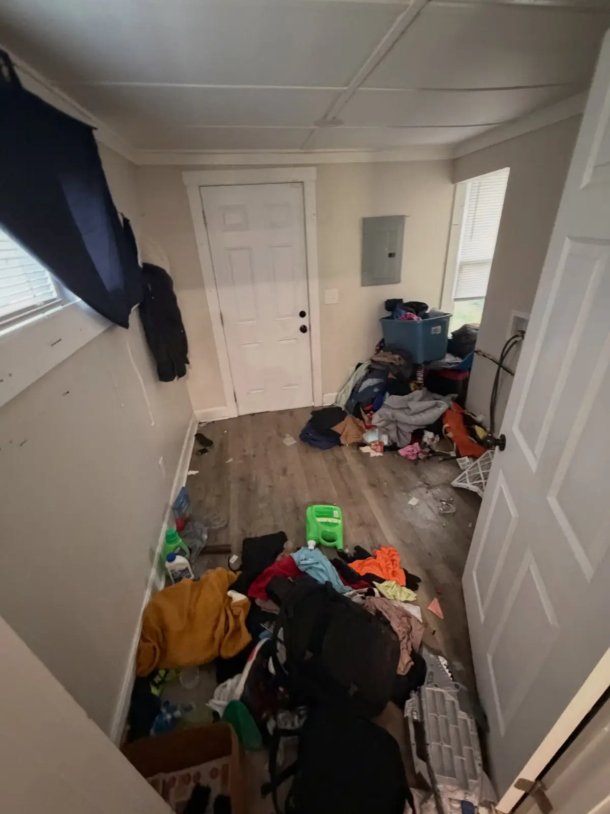 Entry and laundry area of a Mesa home before a cleanout, with clothing, backpacks, storage totes, and household items covering the floor