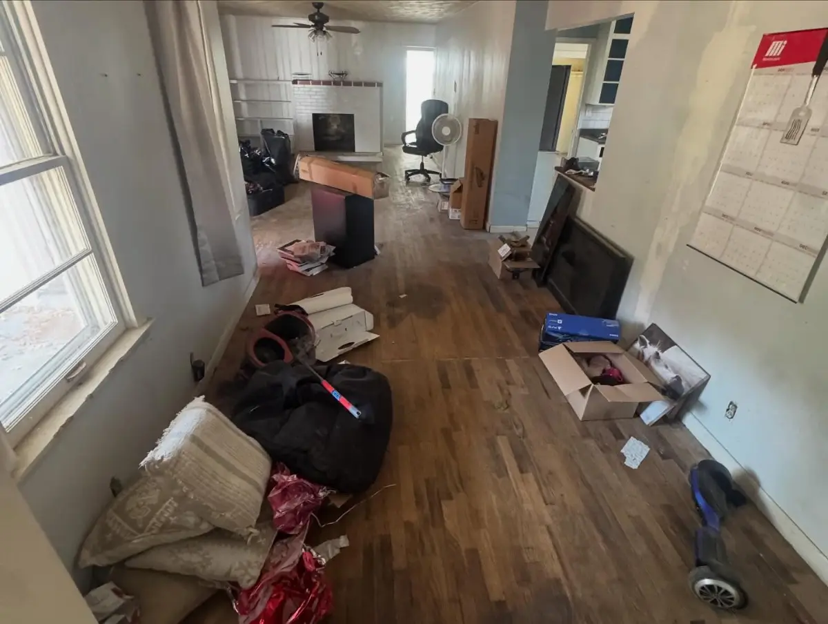 A long hardwood living room in a Mesa house with a desk, a TV stand, bagged items, boxes and loose contents scattered across the floor