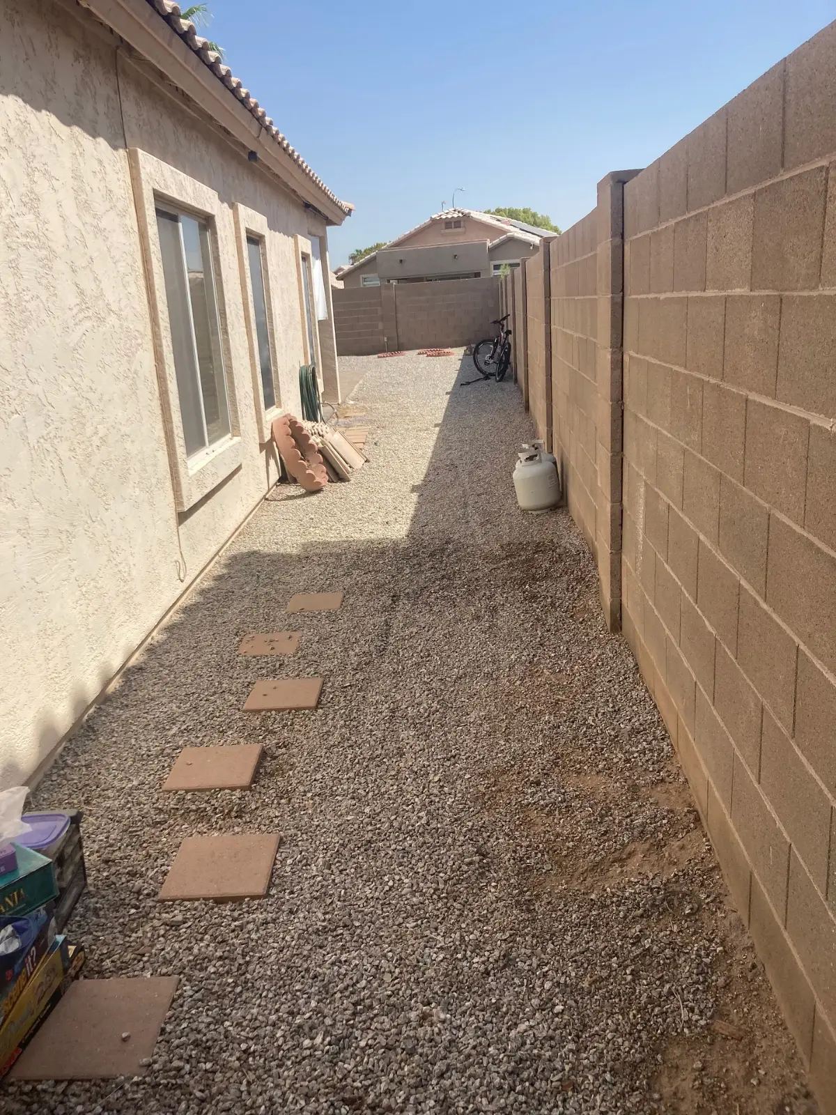 The same Mesa side yard with the gravel path clear; a propane tank and stacked pavers remain by the house