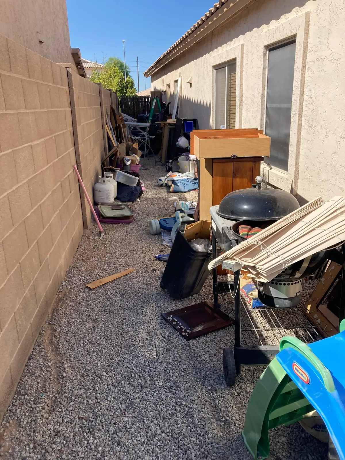 Gravel side yard at a Mesa home crowded with a kettle grill, a wooden cabinet, a plastic playhouse, toys, lumber, tubs and a trash bin