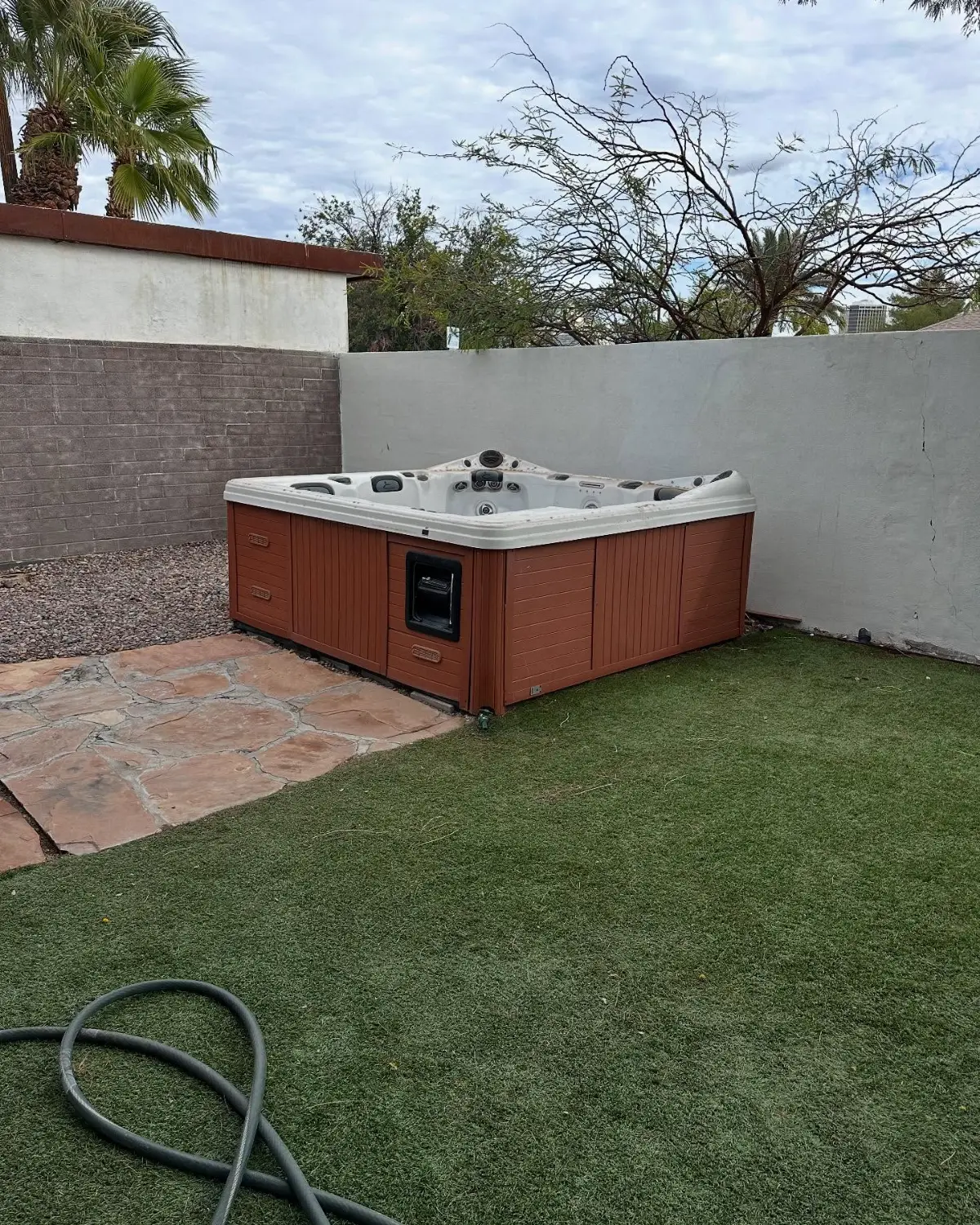 A white hot tub with a terracotta skirt in the corner of a painted block wall on turf and flagstone in a Phoenix backyard