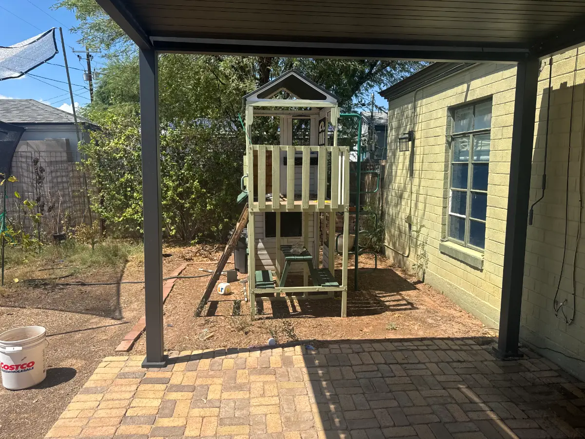 A two-tier wooden playset with a slide standing in a dirt Phoenix backyard under a patio cover beside a green casita