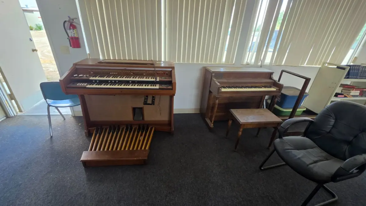 An electric organ, an upright piano, a bench and chairs in a carpeted Sun City room with vertical blinds