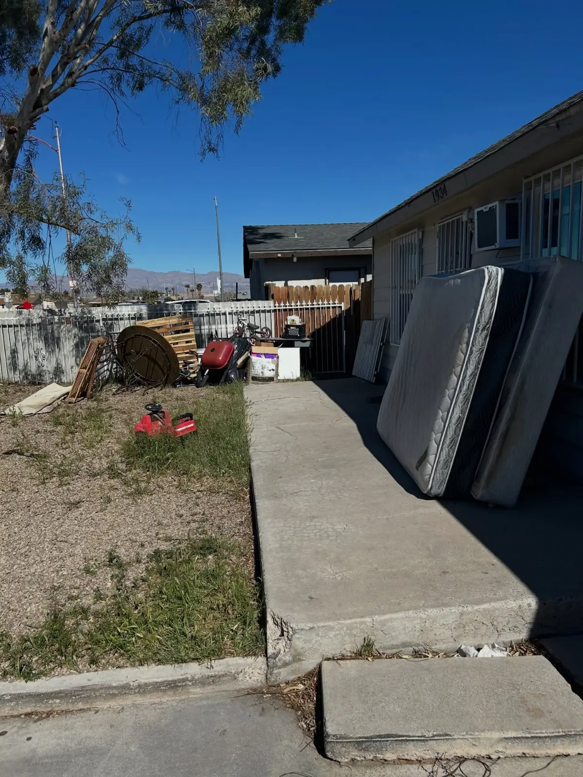 Mattresses and box springs stacked against the side of a Tempe house with a mower and scrap in the yard beyond